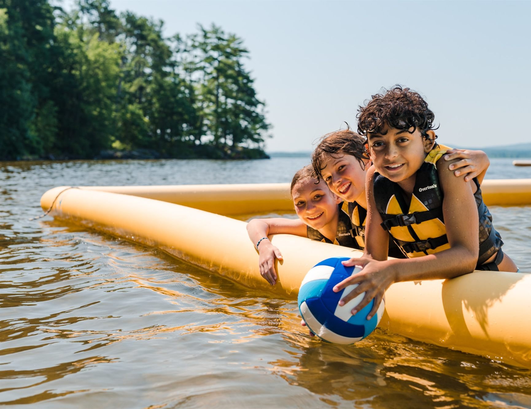 Three campers in life jackets leaning on a floating tube with a volleyball