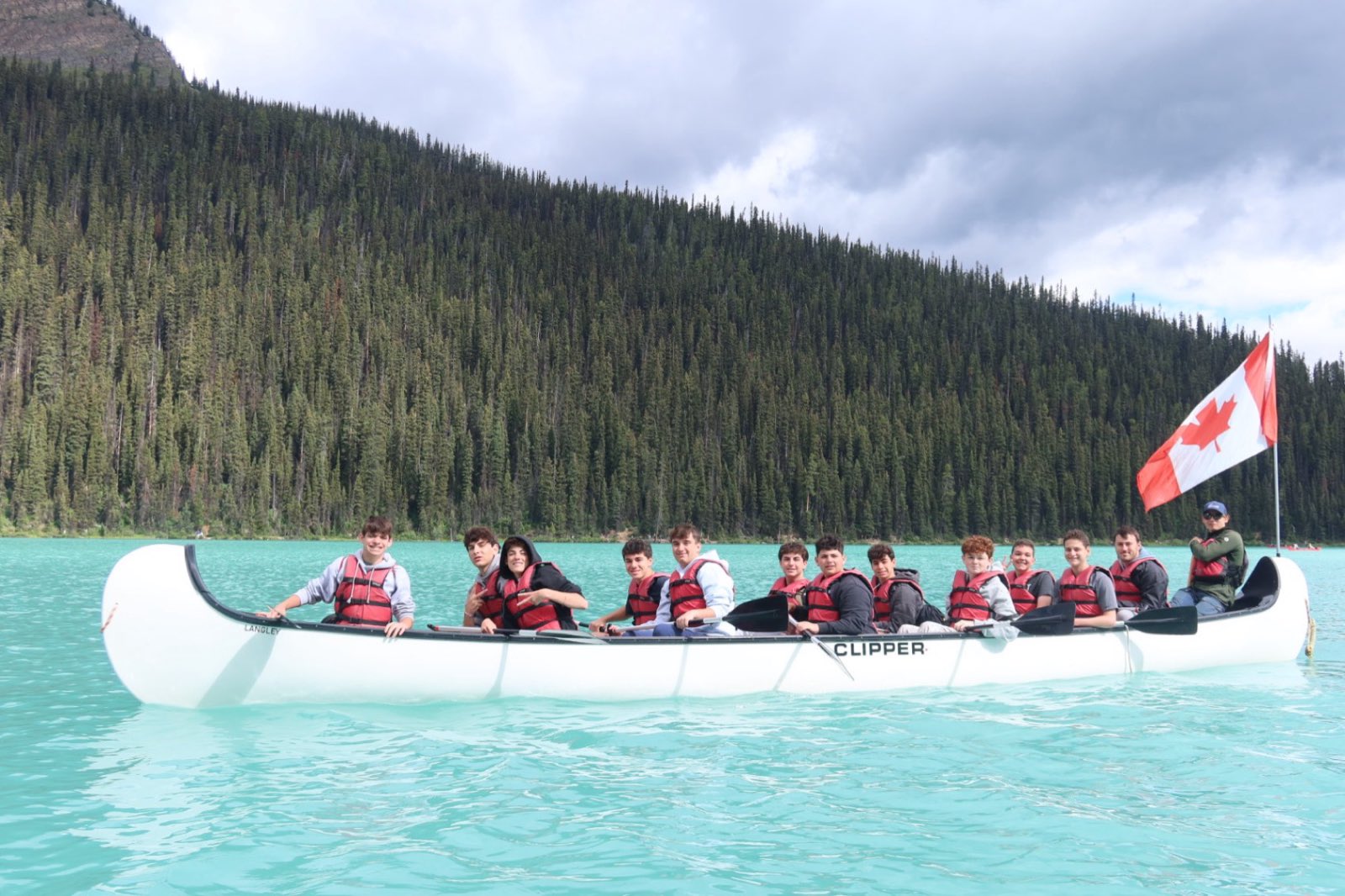 Teens paddling a big canoe flying a Canadian flag across a turquoise mountain lake