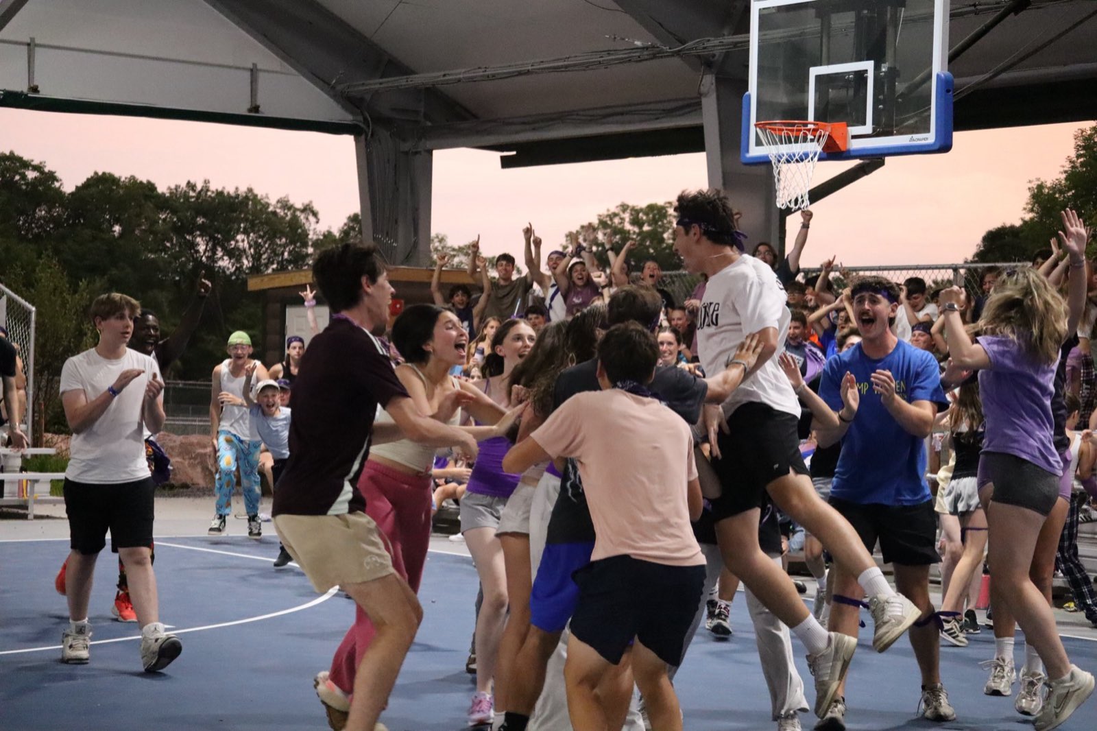 Teen campers mobbing the basketball court at dusk in celebration, one lifted above the crowd