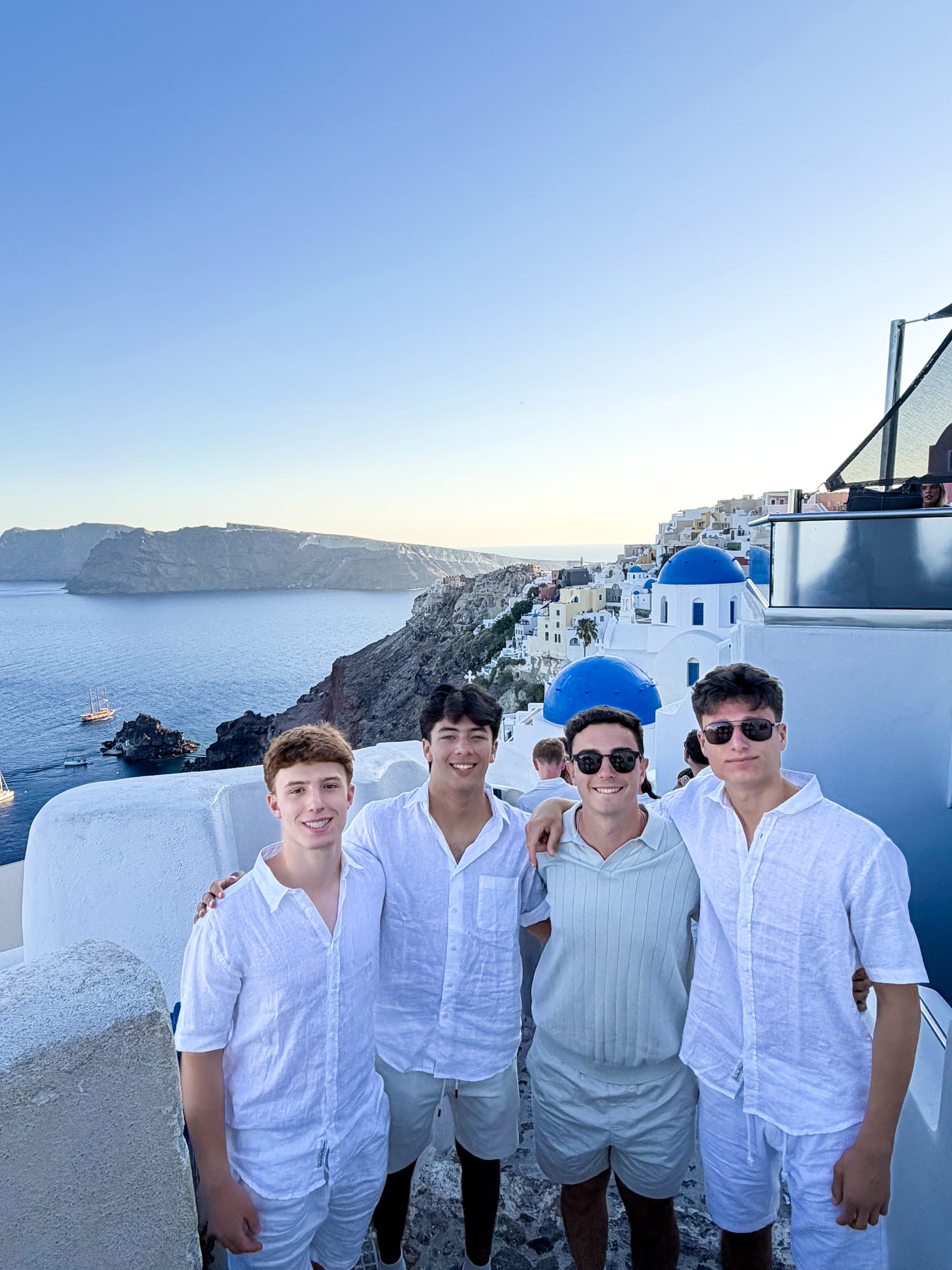 Four teens in white shirts smiling together above the caldera in Santorini