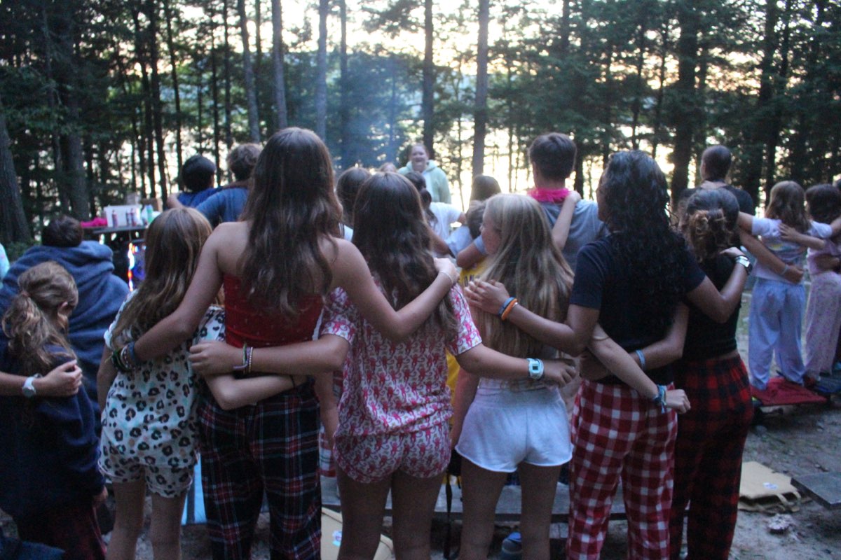 Teen campers standing arm in arm watching the sunset through the pines