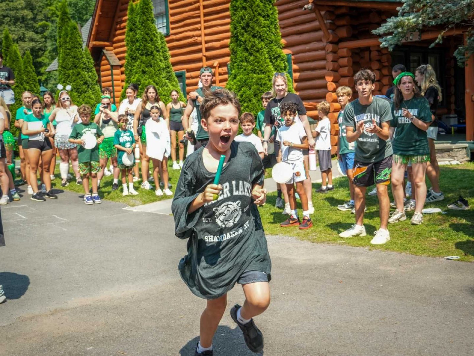 A camper races toward the camera cheering, teammates in green lining the path outside a log lodge